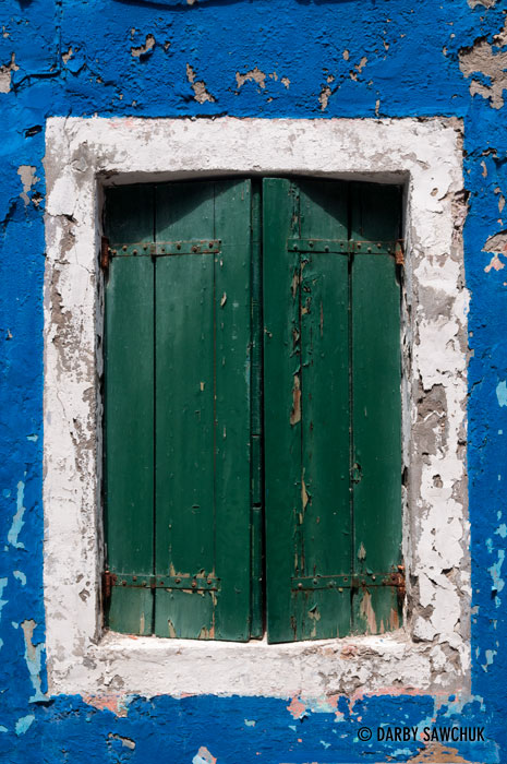 Paint flakes away from around a window of a bright blue house in Burano, Italy.