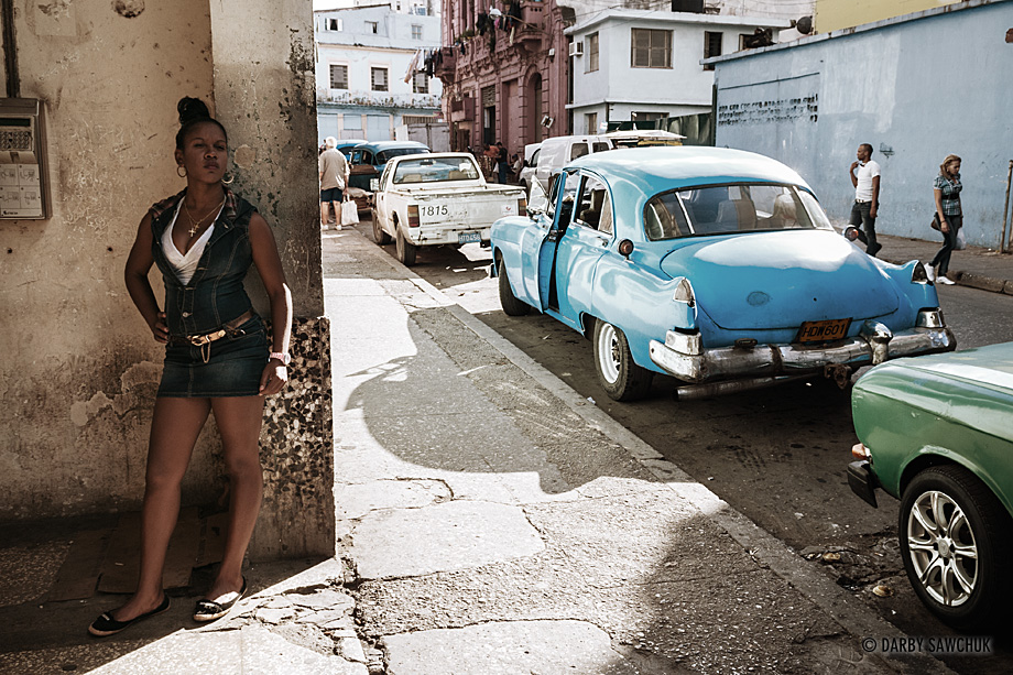A woman waits in the shade with vintage american cars in the background in central Havana.