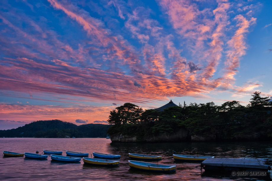 Rowboats float in Matsushima Bay with Godaido Hall in the background at sunset.