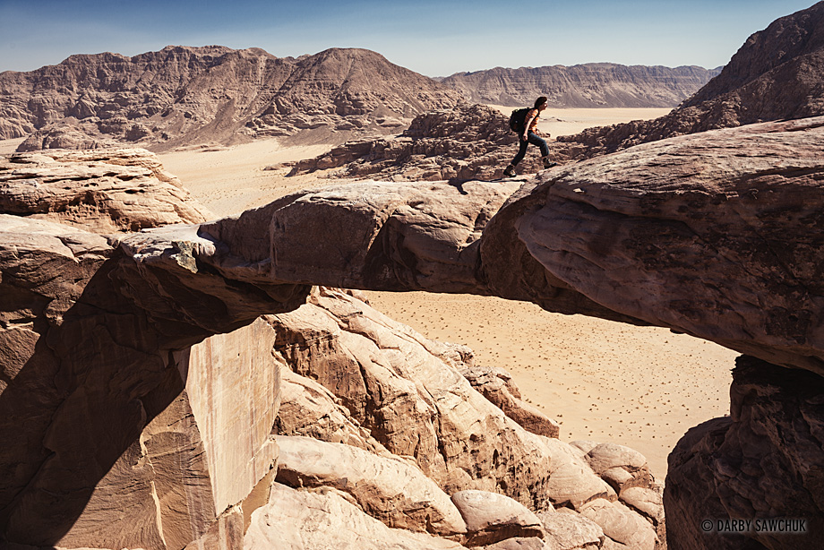 A hiker walks across the narrow rock bridge of Burdah Arch high above Jebel Burdah and the desert of Wadi Rum.