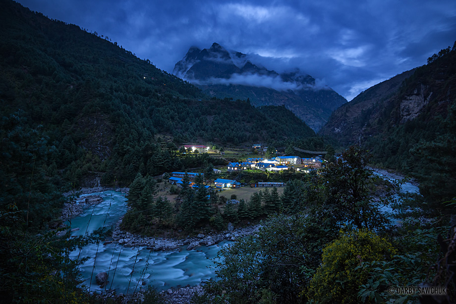 The Dhudh Kosi river winds past guesthouses lit up during a quiet evening in Phakding with cloud-enshrouded Himalayan mountains behind.