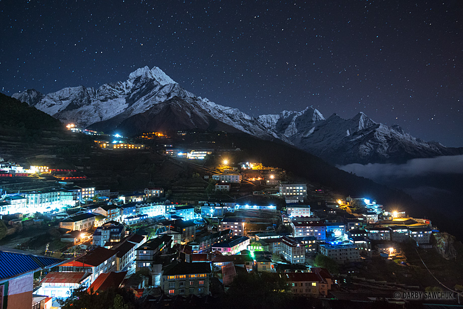 Stars shine at night over Namche Bazaar and the moonlit Himalayan mountains in the background.