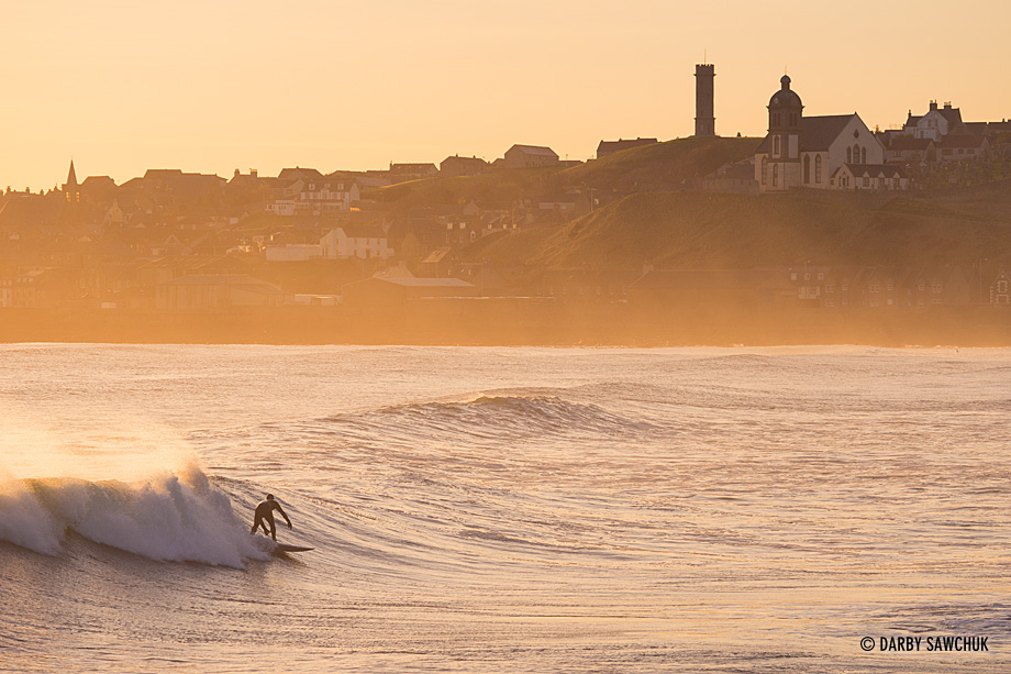 A surfer braves the cold North Sea waters at sunrise in Banff Bay with MacDuff in the background.