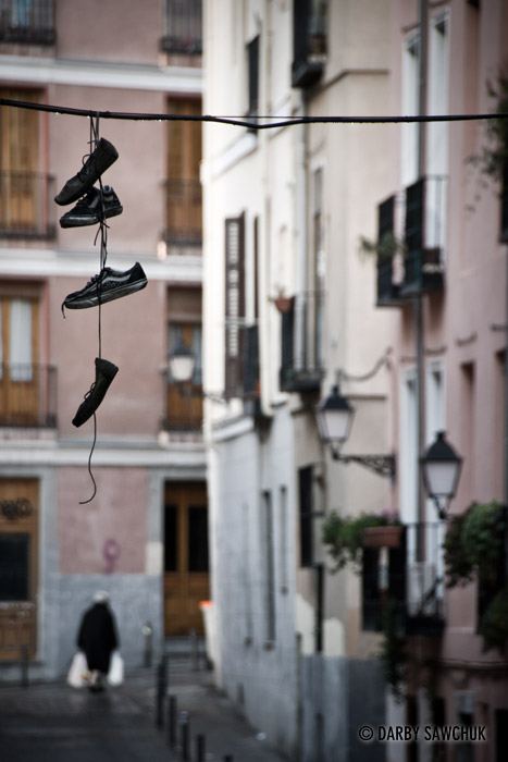 Shoes dangle from a wire while a woman carries her shopping in the distance in a back street of Madrid, Spain.