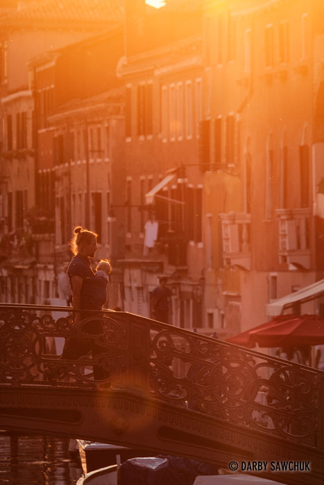 A man carries a baby across a bridge in the Cannaregio district at sunset.