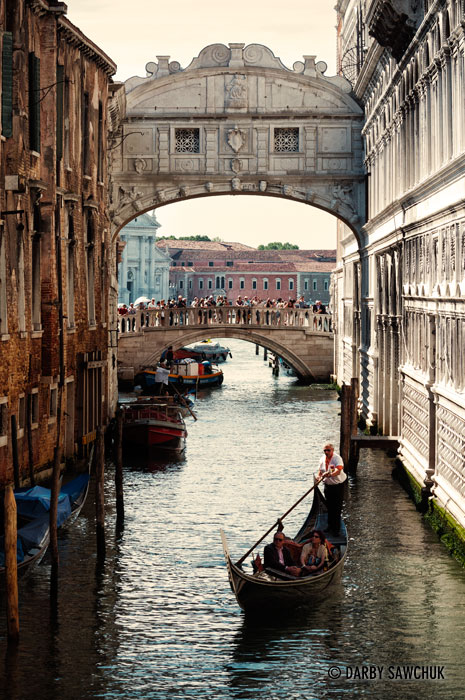 A gondola glides under the Bridge of Sighs while tourists admire the view in the background.
